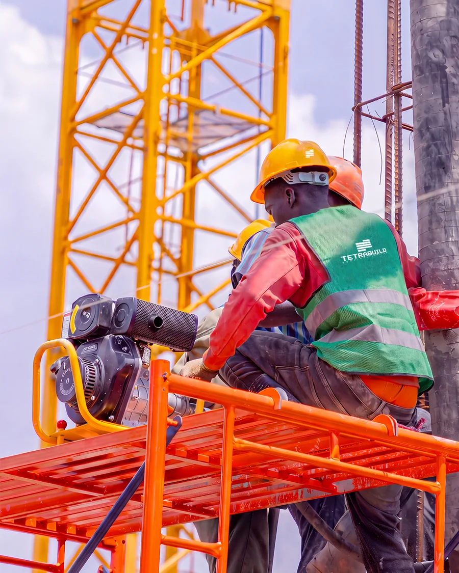 Two Tetrabuild workers in branded hi-vis on an orange scaffold platform, working on a reinforced concrete column beneath a tower crane