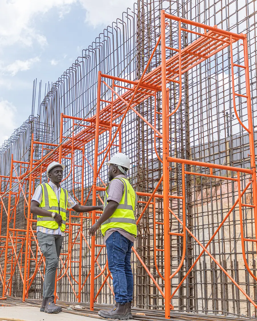 Two Tetrabuild engineers in hi-vis and hard hats reviewing work in front of H-frame scaffolding against a reinforced concrete retaining wall